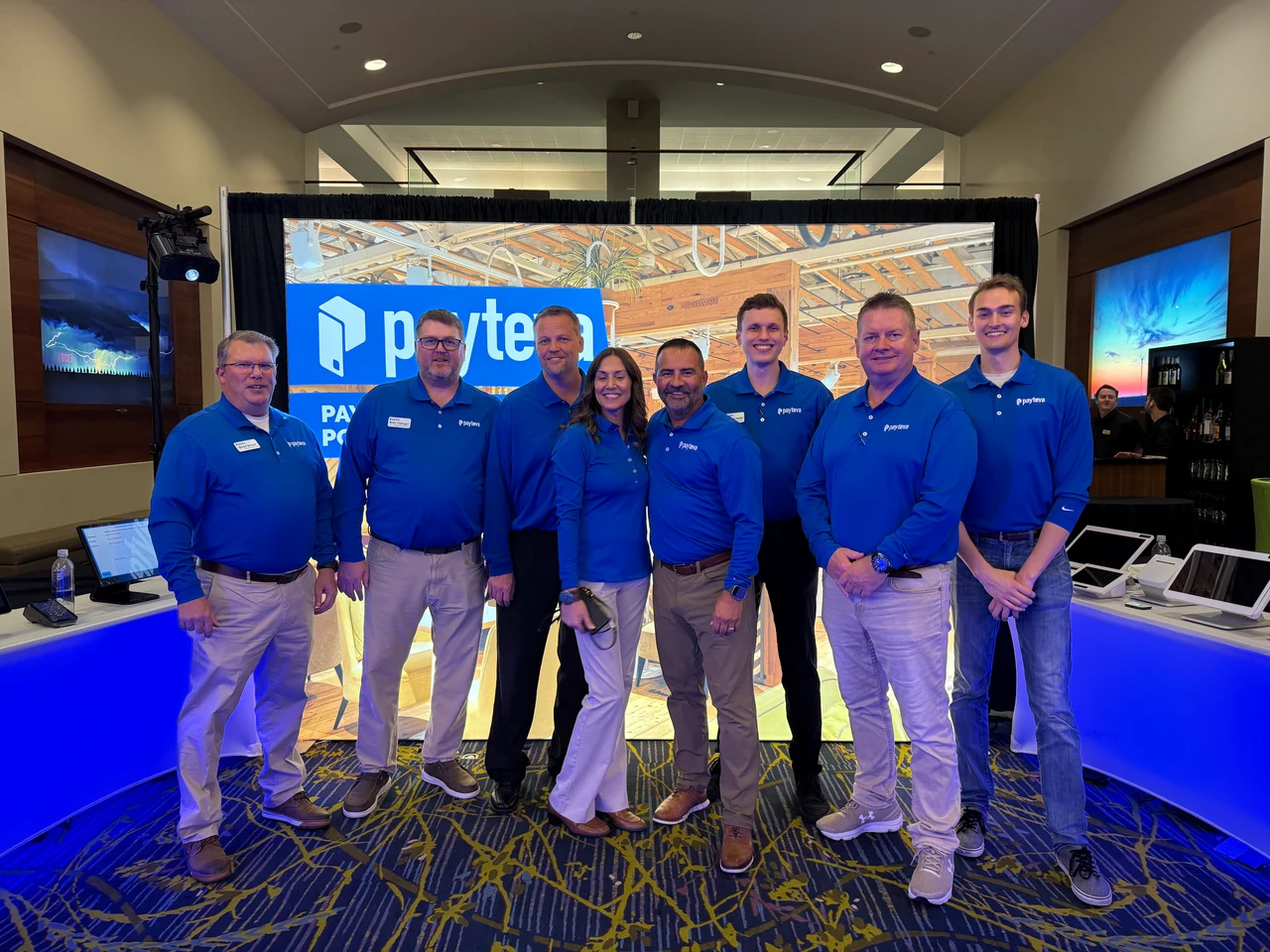 Eight Payteva representatives in matching blue Payteva shirts standing together at an industry event